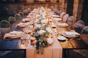 in backyard of villa in Tuscany there is banquet wooden table decorated with cotton and eucalyptus compositions, glasses, burning candles and plates are placed on table, evening